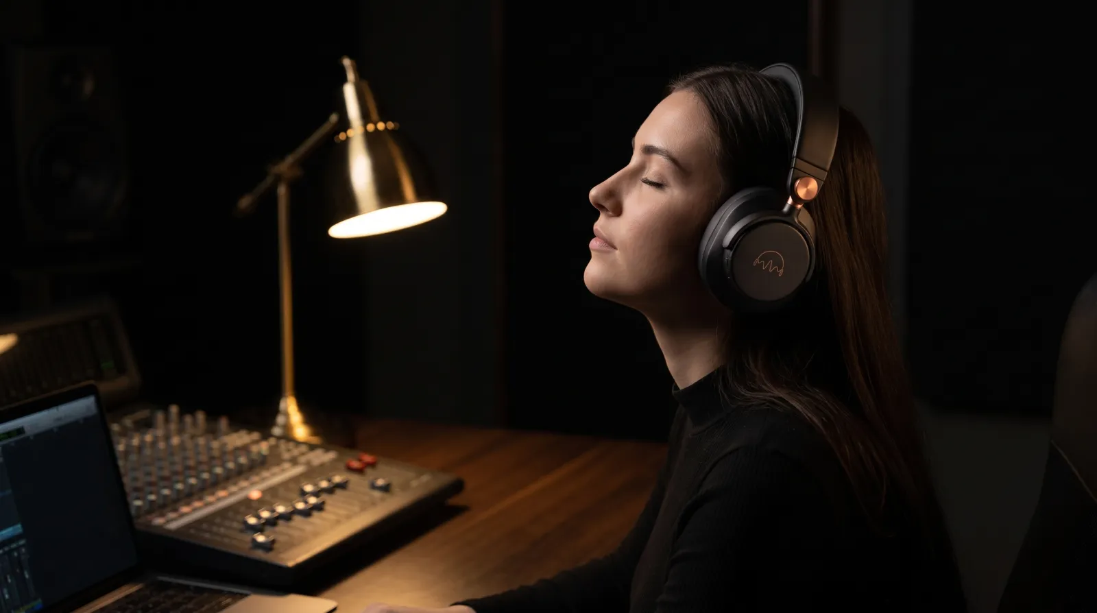 A woman at a desk lamp at night wearing the A1 headphones, eyes closed, beside a mixing desk